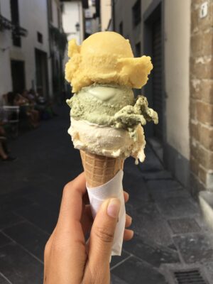 someone holding a cone with three scoops of gelato that are yellow, green, and tan with an Italian alley in the background