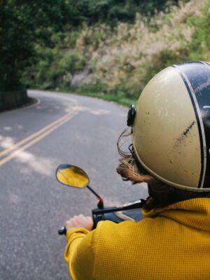 A photo taken from the back of a vespa showing the driver in yellow and a helmet and the grass-lined road in front