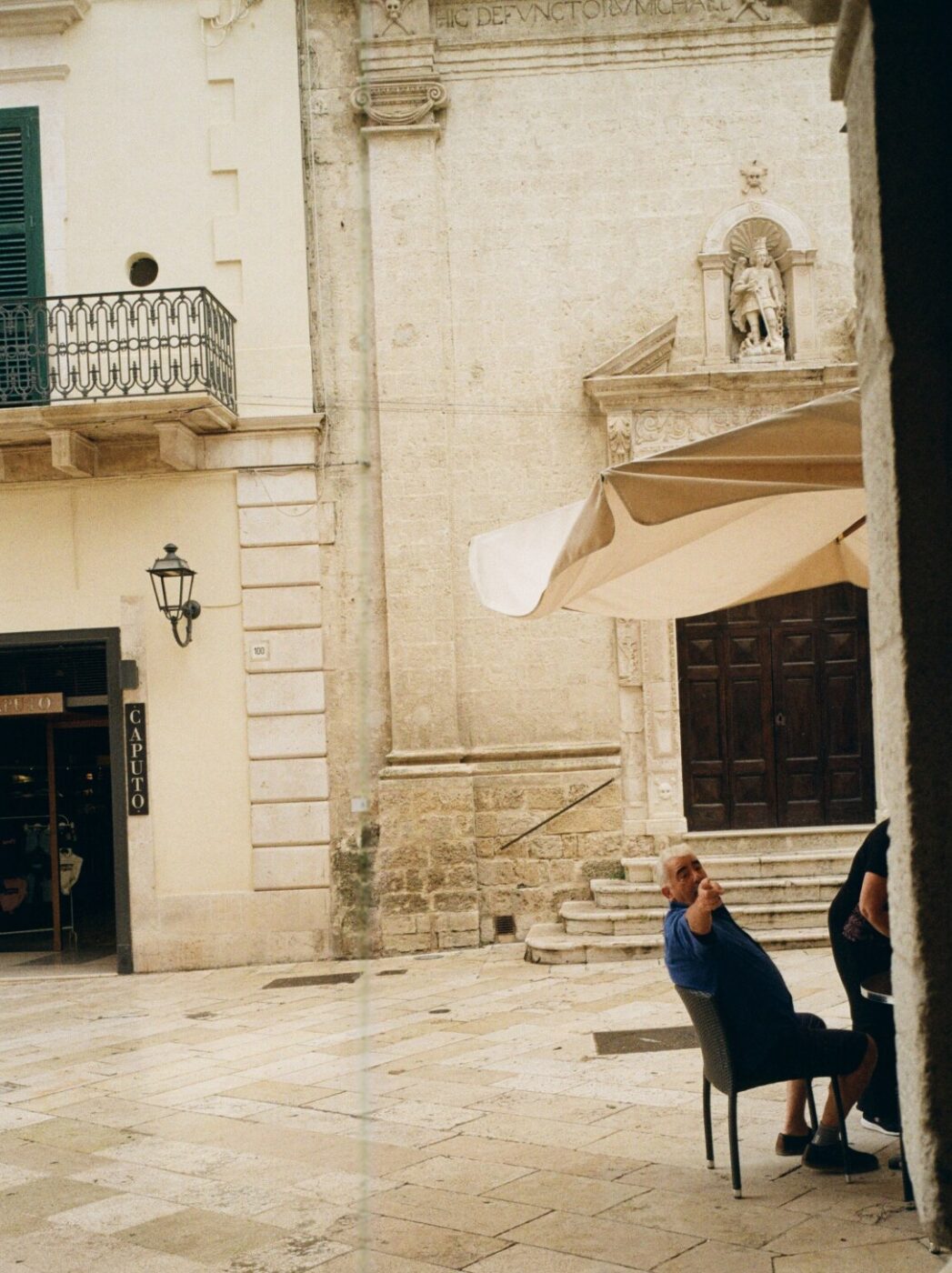 a vintage photo of a piazza in Altamura and a man sitting at a cafe and pointing towards the camera