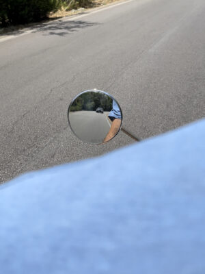 A photo taken from the back of a vespa showing a tiny, circular side mirror with a small car in the background and a hint of the driver's tan arm with a blue short sleeved shirt