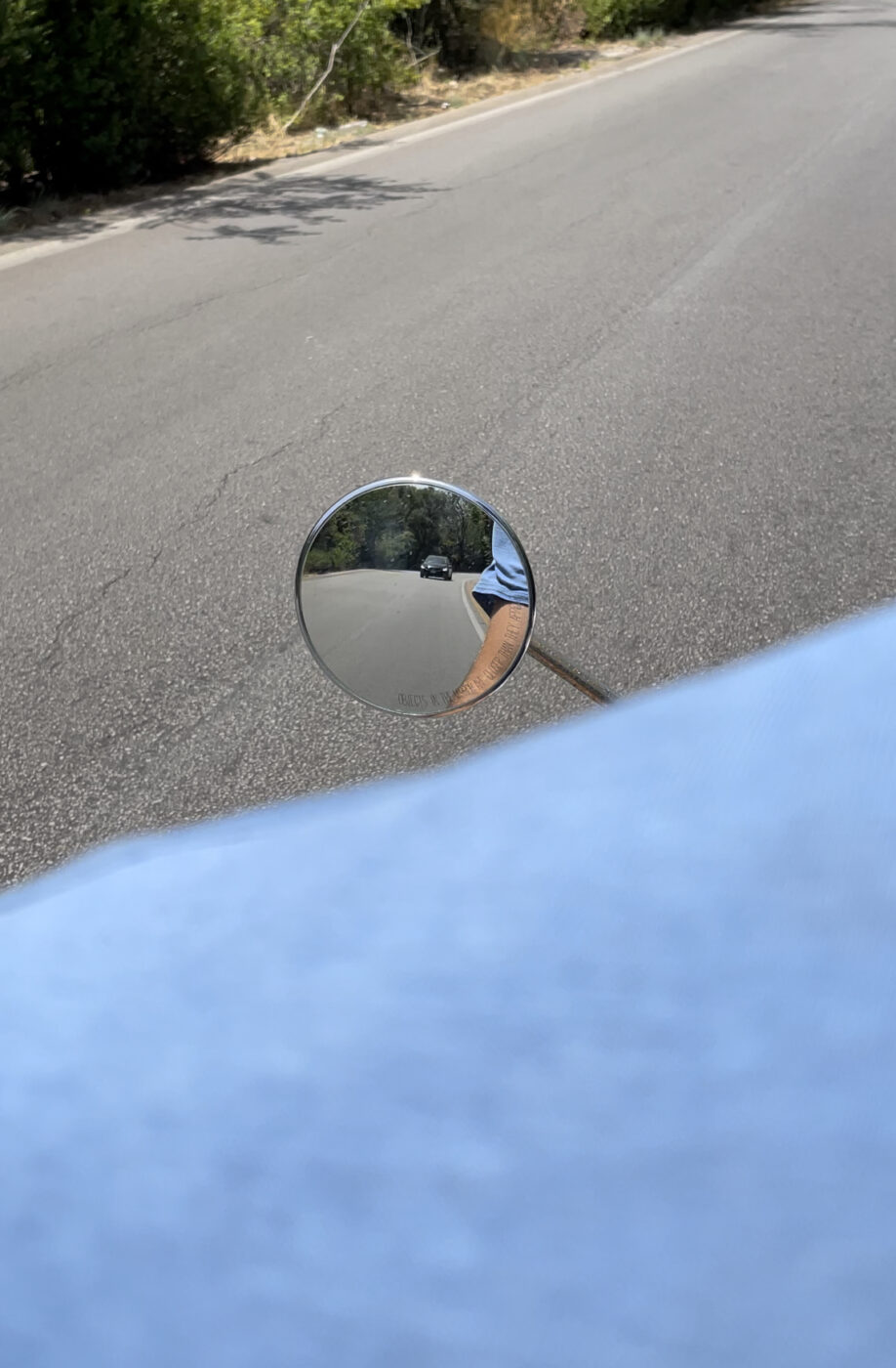A photo taken from the back of a vespa showing a tiny, circular side mirror with a small car in the background and a hint of the driver's tan arm with a blue short sleeved shirt