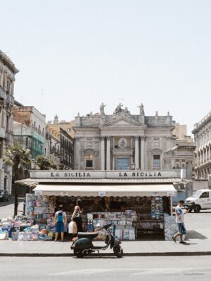 a photo of a newsstand called "La Sicilia" in a piazza in Sicily with a vespa parked out front