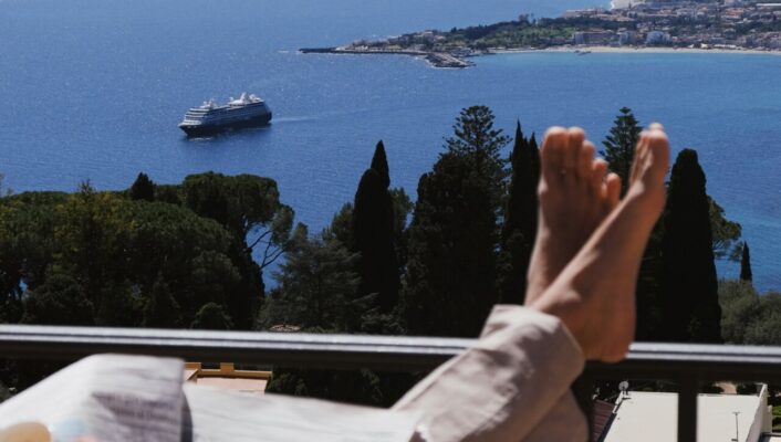 a photo of a woman lounging, drinking coffee, and reading a newspaper on a balcony overlooking the sea in Sicily