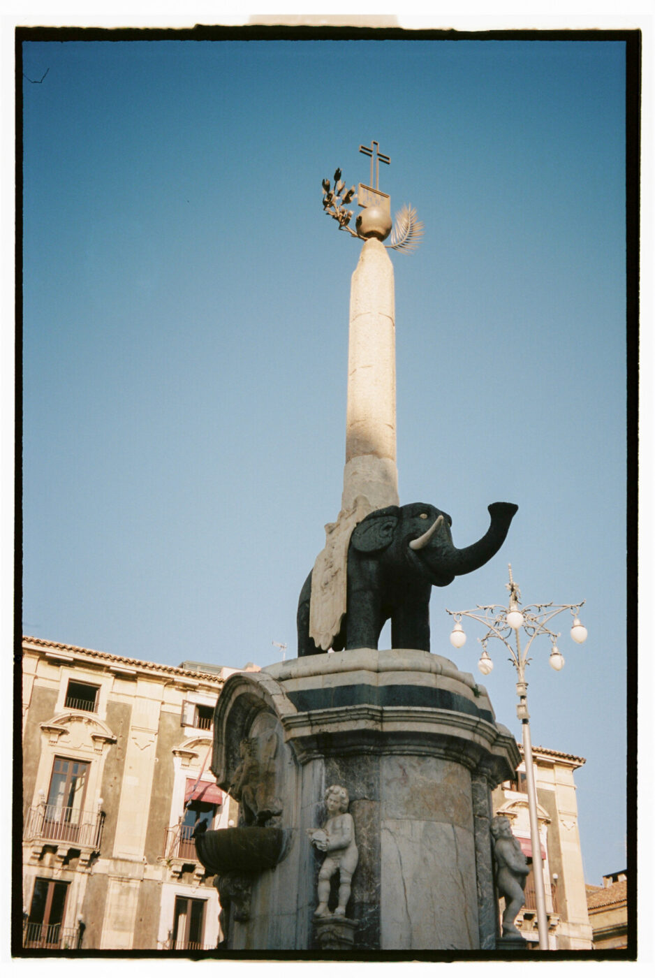 Photography by Gina Spinelli of a statue of an elephant with an obelisk coming out of it topped with a cross in the center of a piazza in Catania