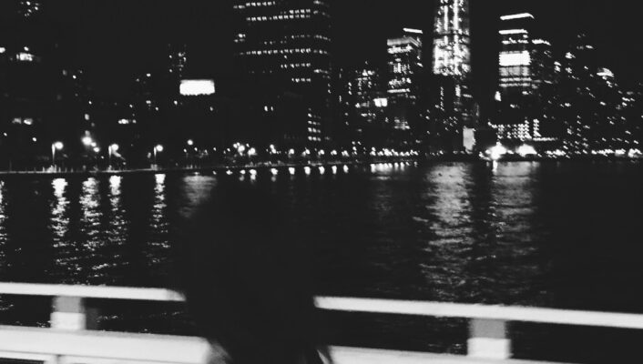 A black and white photo of a woman in the foreground and the Freedom Tower at Battery Park in NYC at night across the river