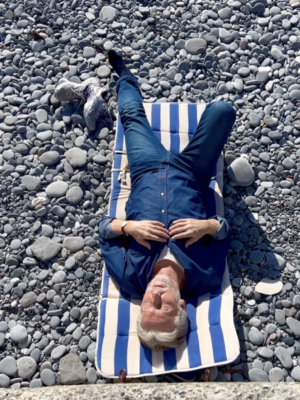 A photo taken from above of a man in navy blue lying on a blue and white striped towel on a gray rocky beach in Genova