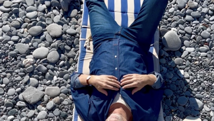 A photo taken from above of a man in navy blue lying on a blue and white striped towel on a gray rocky beach in Genova