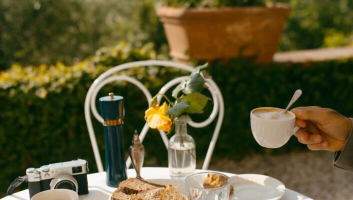 A sunlit table with plates, bread, coffee cups, and glasses; a hand lifts a cup. Camera, vase with yellow rose, red flowers behind.