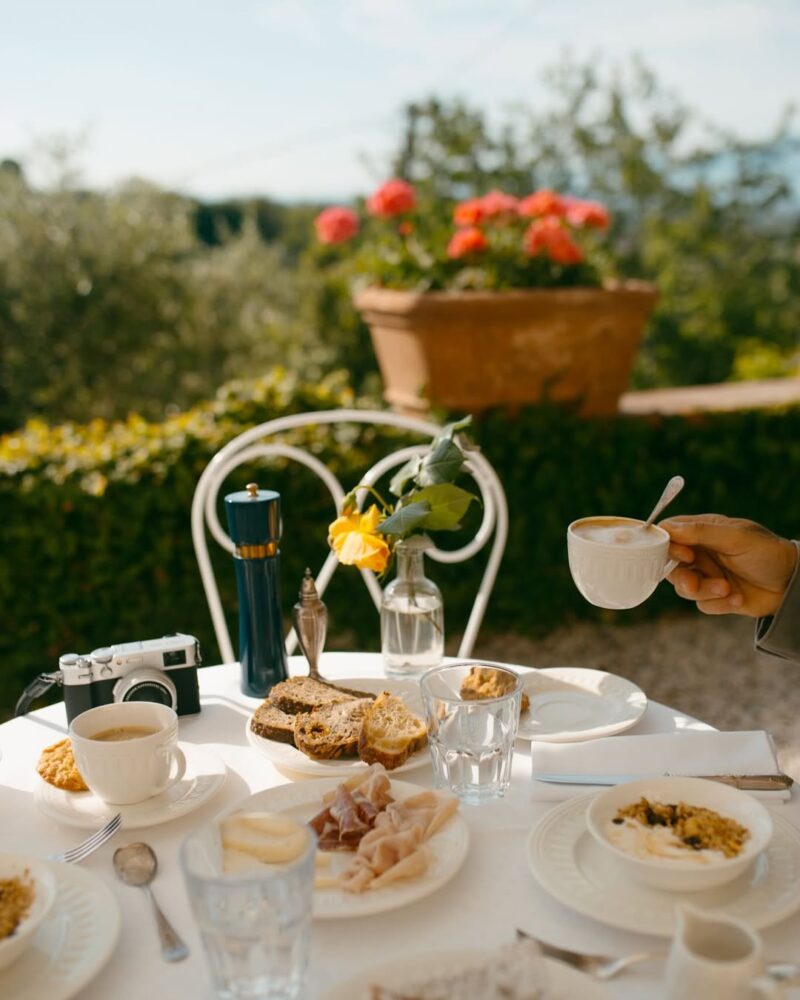 A sunlit table with plates, bread, coffee cups, and glasses; a hand lifts a cup. Camera, vase with yellow rose, red flowers behind.