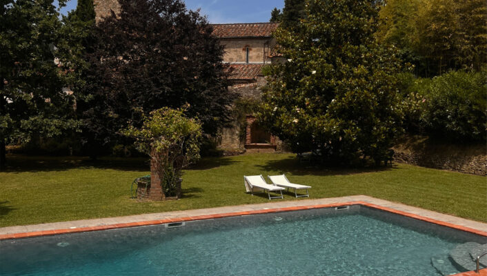 A rectangular pool with grass and two lounge chairs; a stone building with a tower and trees under a blue sky with clouds.