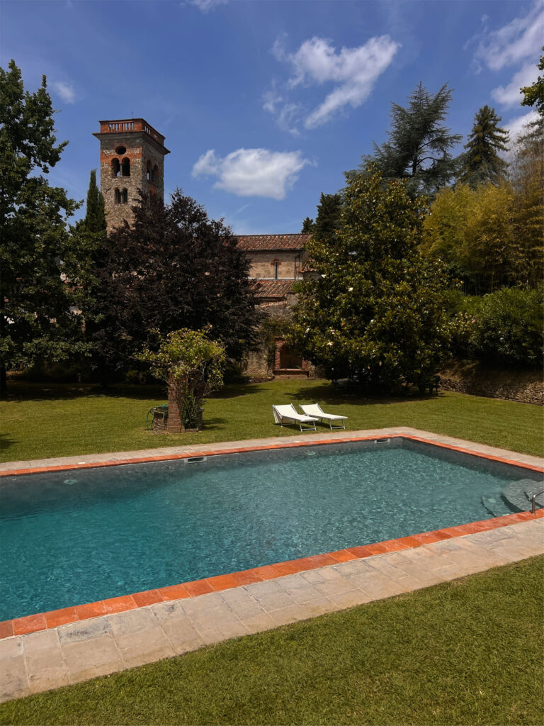 A rectangular pool with grass and two lounge chairs; a stone building with a tower and trees under a blue sky with clouds.