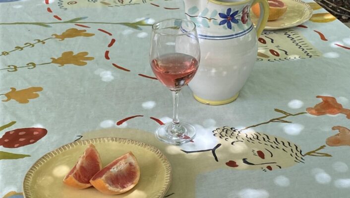 ceramics by Gabriella Picone: a shaded outdoor table with a light blue tablecloth with a woman on it, two wine glasses with juice, a ceramic white pitcher, and two yellow ceramic plates with slices of grapefruit