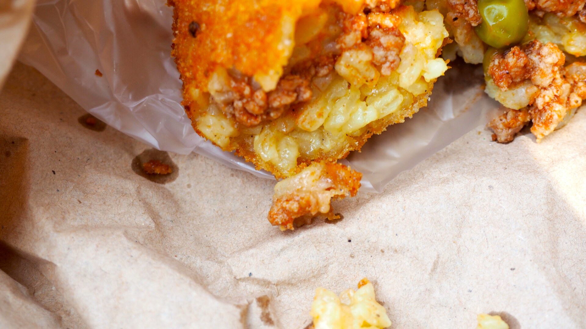 a half eaten arancino with the meat, cheese, and pea filling spilling out of the crunchy shell on oily wax paper
