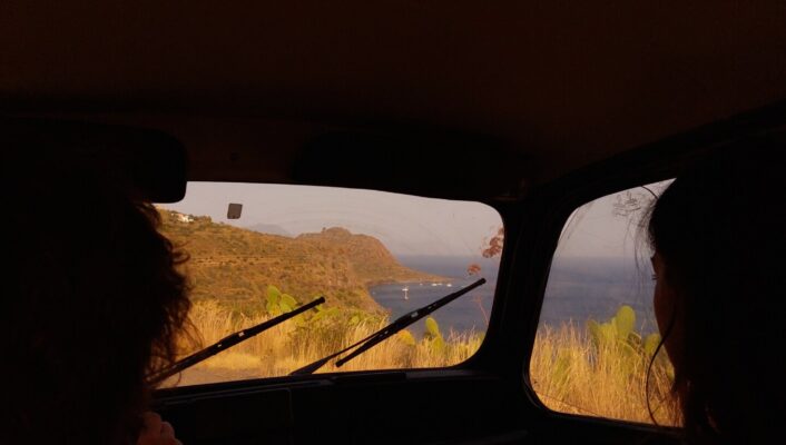A photo taken from the backseat of a car of a of a couple in the front seats with the island of Filicudi out the window