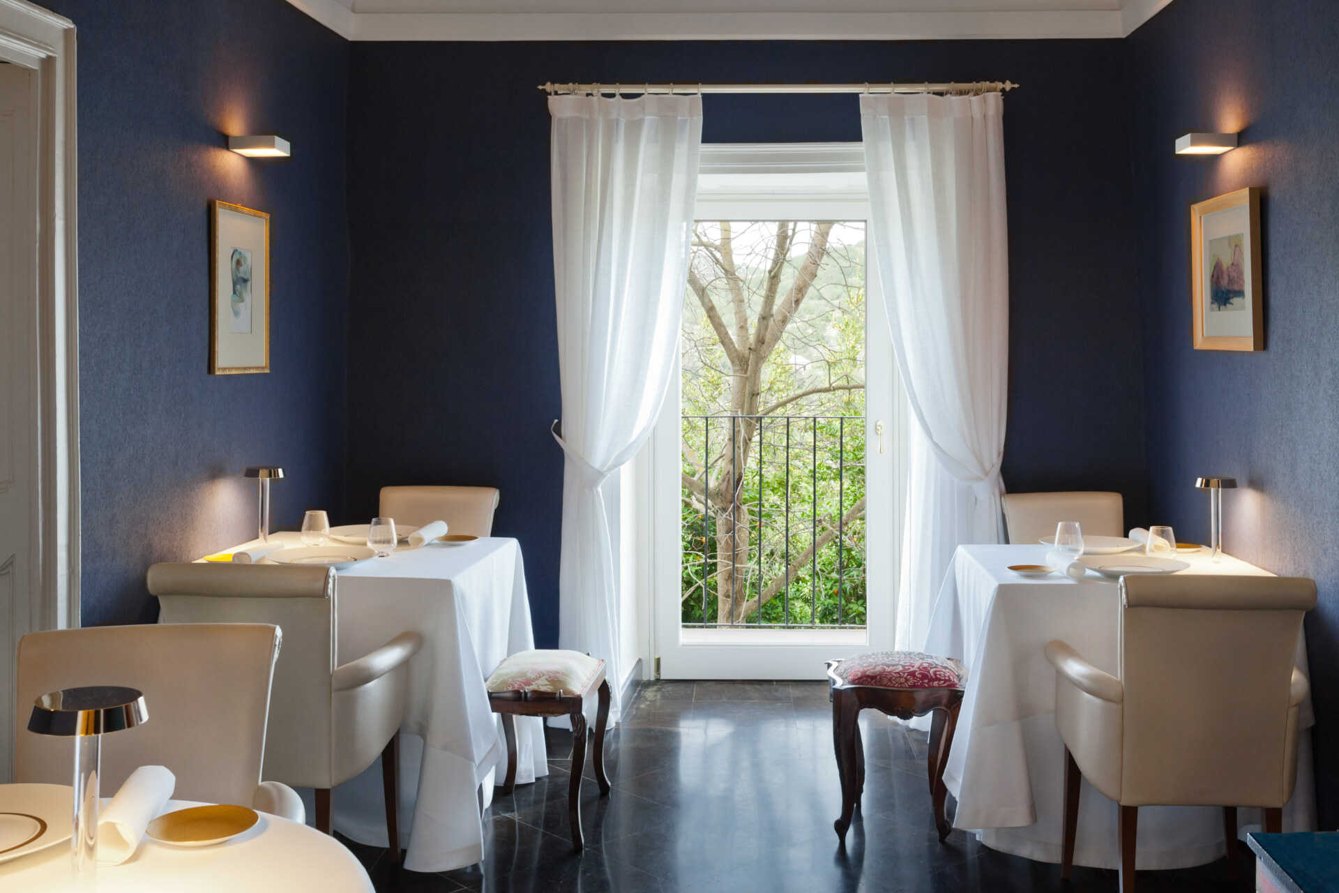 an interior of Duomo restaurant; navy walls, a window overlooking green trees, and three 2 person white tables set elegantly© Benedetto Tarantino