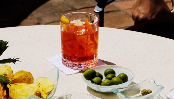 am elegant photo of a table at aperitivo with green olives, peanuts, an orange spritz in a highball glass, and potato chips