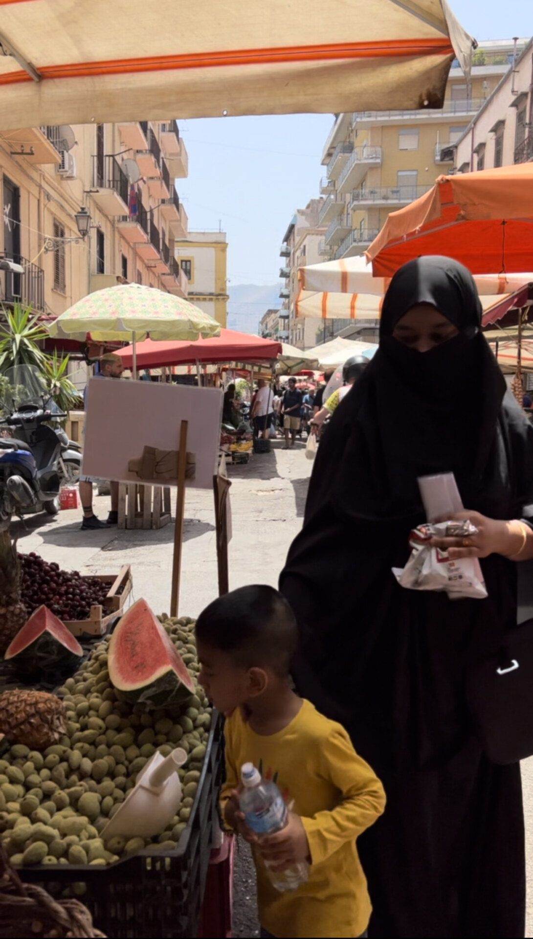 a mother and son shopping at palermo's Ballarò market; the son is looking at containers of grapes and watermelon