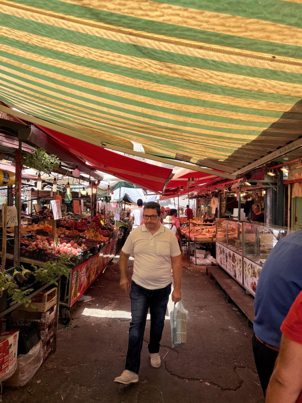 a man walking through a shaded portion of palermo's Ballarò market with stands of fruit and vegetables on either side