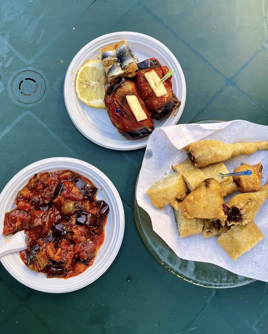 three dishes on disposable plates on a aqua table: caponata, assorted fried things, and anchovies wrapped around something on a stick and eggplant with cheese and sauce on a stick
