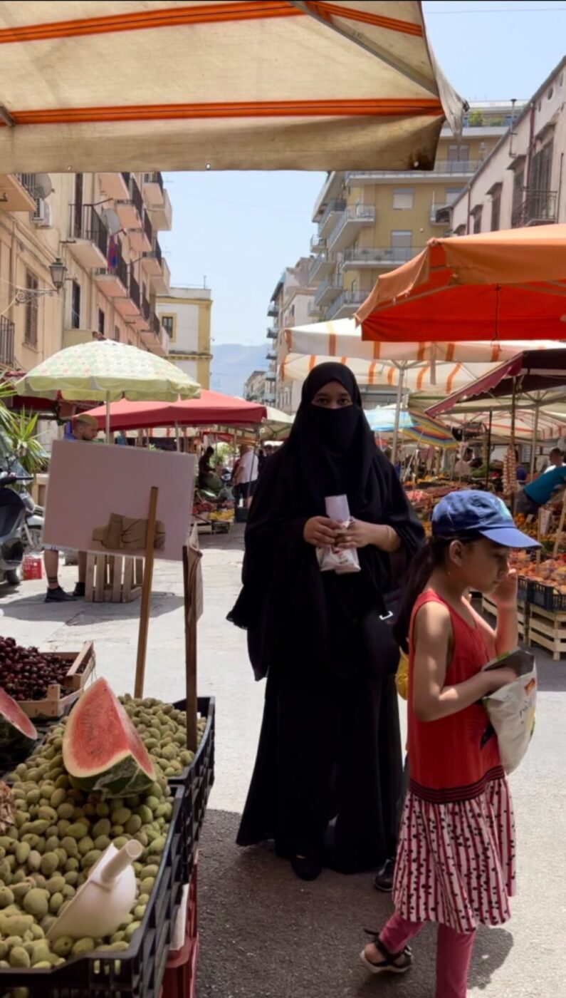a woman wearing a black chador walking through Palermo's Ballaro market while a yoing girl in front eats chips from a bag