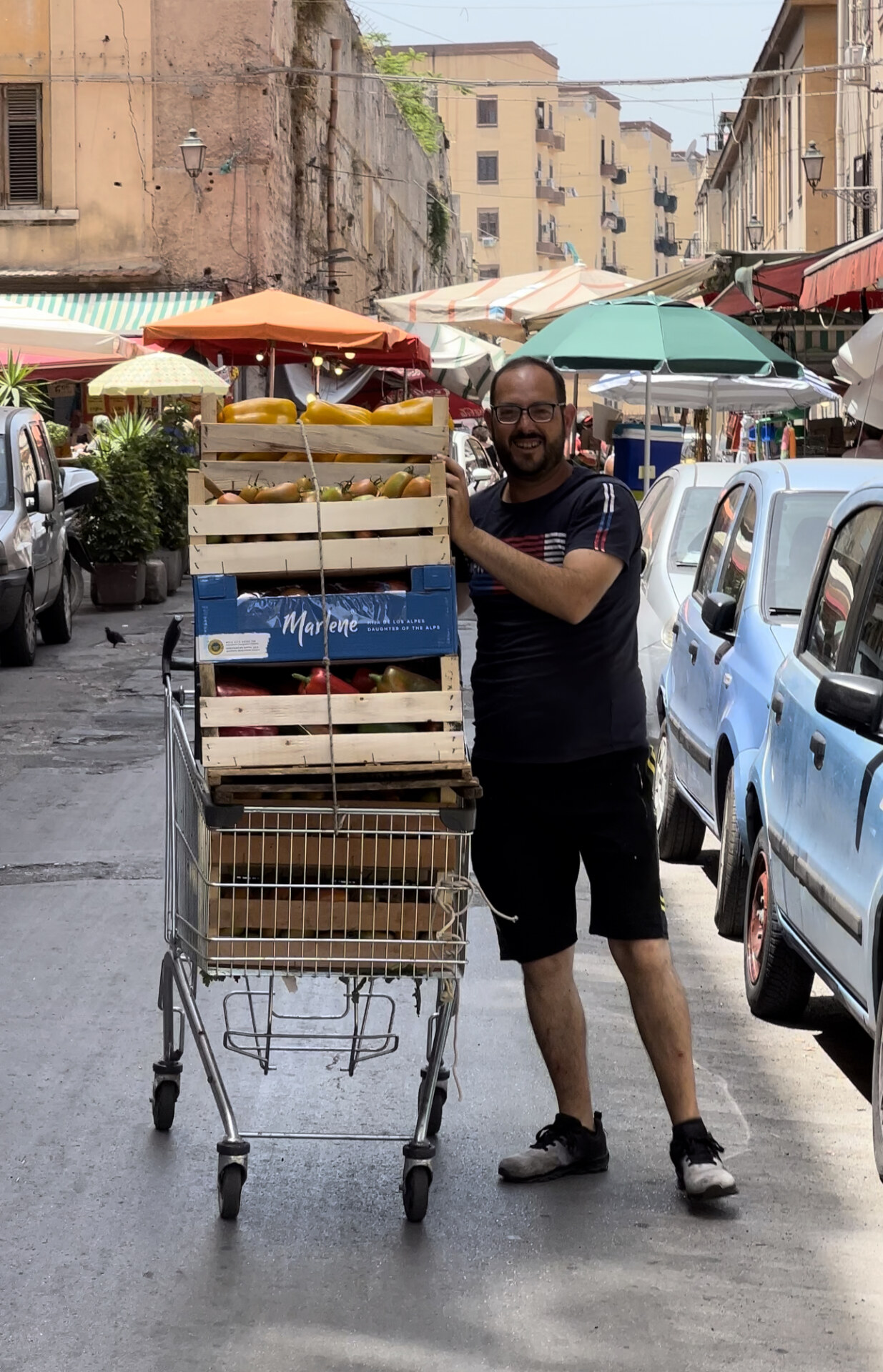 A man pushing a shopping cart filled with containers of vegetables and fruit to sell at Palermo's Ballarò market