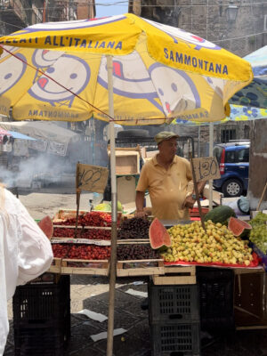 A man selling fruit under a yellow sammontana umbrella with smiley faces at Palermo's Ballarò market