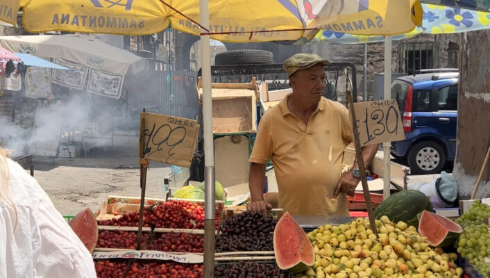 A man selling fruit under a yellow sammontana umbrella with smiley faces at Palermo's Ballarò market