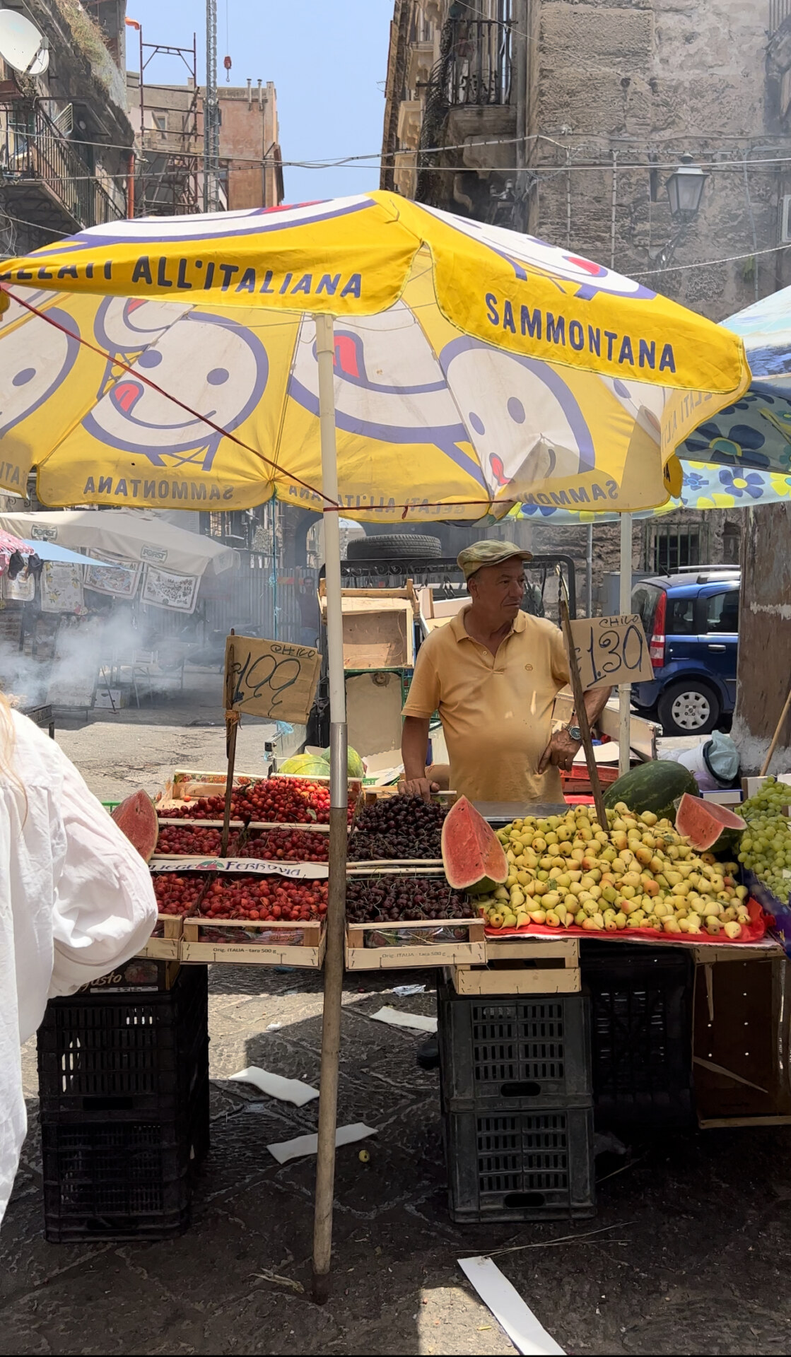 A man selling fruit under a yellow sammontana umbrella with smiley faces at Palermo's Ballarò market