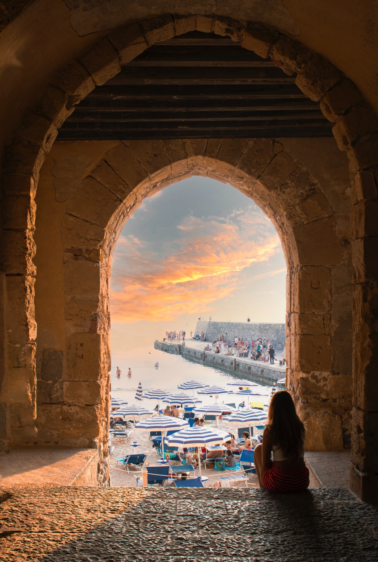 a girl sitting in an archway looking out onto the beach with stripped umbrellas at sunset of Cefalu, Sicily