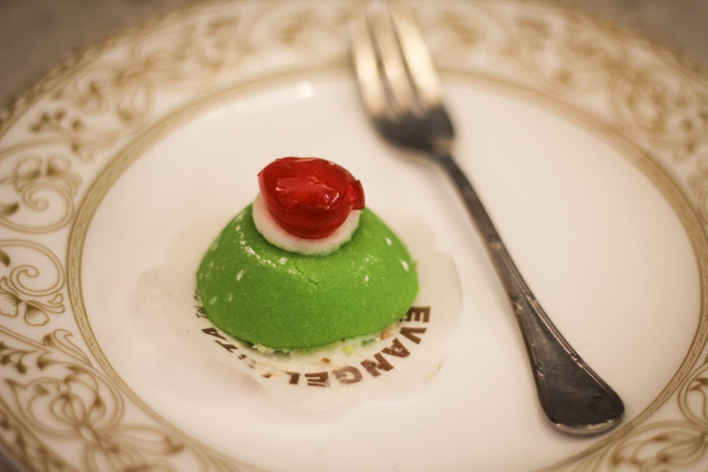 A bright green, round cassata with a bright red cherry sitting on a pretty plate with a fork next to it