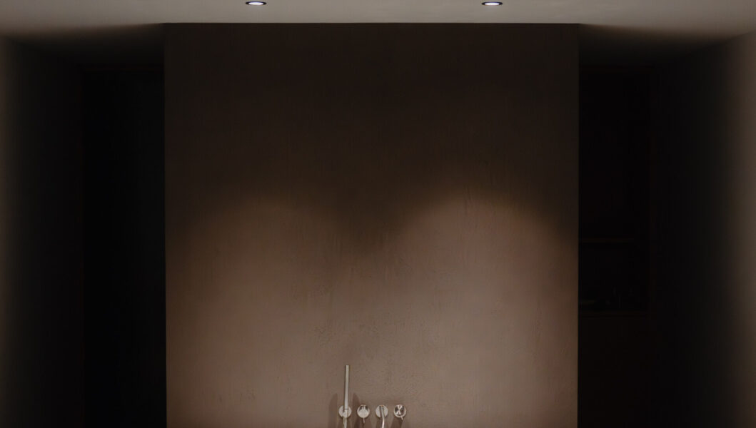 A minimal bathroom with a white tub under two lights, wooden stool with towels and bottle, all in neutral tones, peaceful vibe.