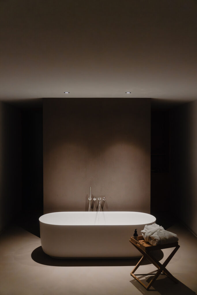 A minimal bathroom with a white tub under two lights, wooden stool with towels and bottle, all in neutral tones, peaceful vibe.