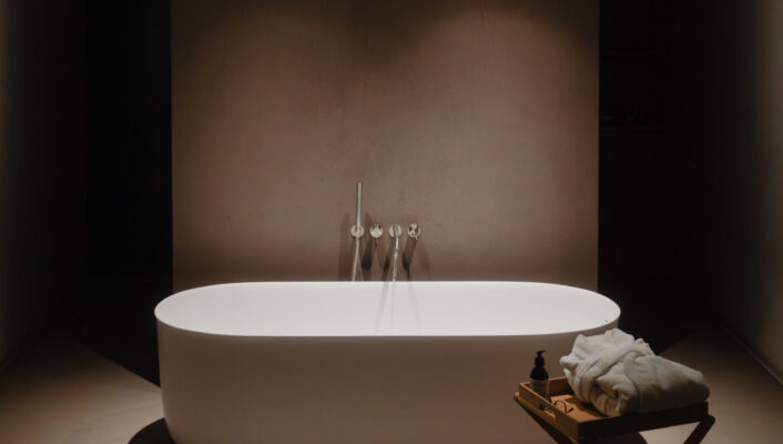 A minimal bathroom with a white tub under two lights, wooden stool with towels and bottle, all in neutral tones, peaceful vibe.