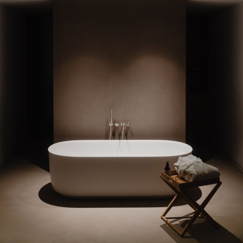 A minimal bathroom with a white tub under two lights, wooden stool with towels and bottle, all in neutral tones, peaceful vibe.