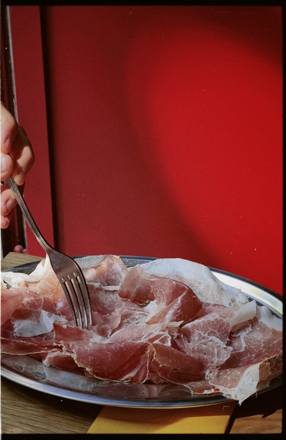 someone holdng a fork over a silver platter filled with prosciutto in front of a red wall