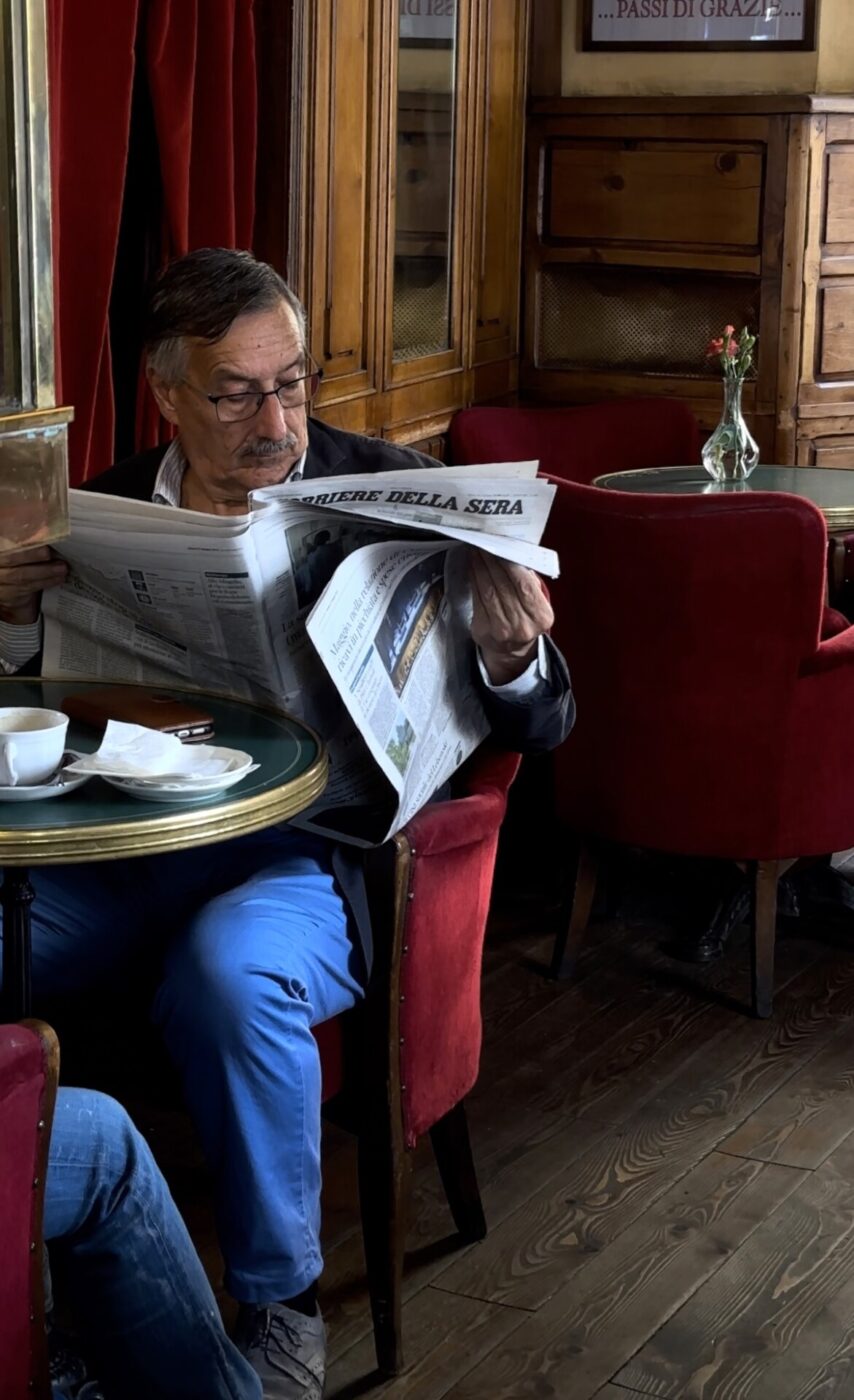 a man with glasses and a mustache sitting in a red velvet chair in a dark green room reading a newspaper