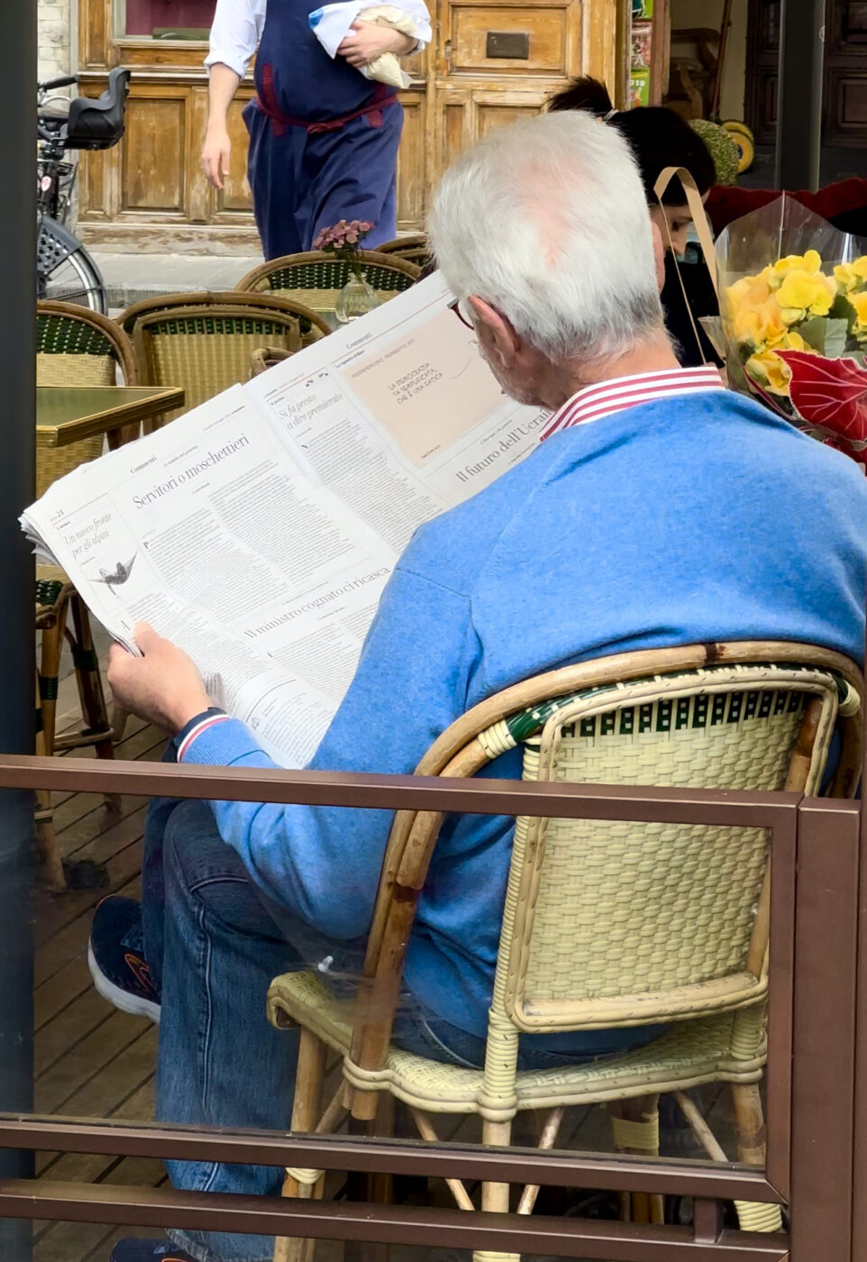 a man in a blue shirt shown from the back sitting at a caffe and reading a newspaper