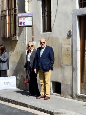 an older Italian couple waiting for the bus in Florence - both are dressed in blazers and pants, wearing sunglasses, and looking past the camera