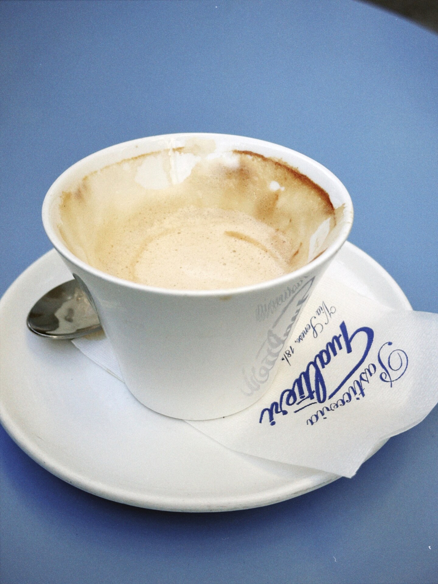 a half-drank espresso in a white cup on a light blue table from Pasticceria Gualtieri with a napkin bearing the pasticceria's name in navy cursive