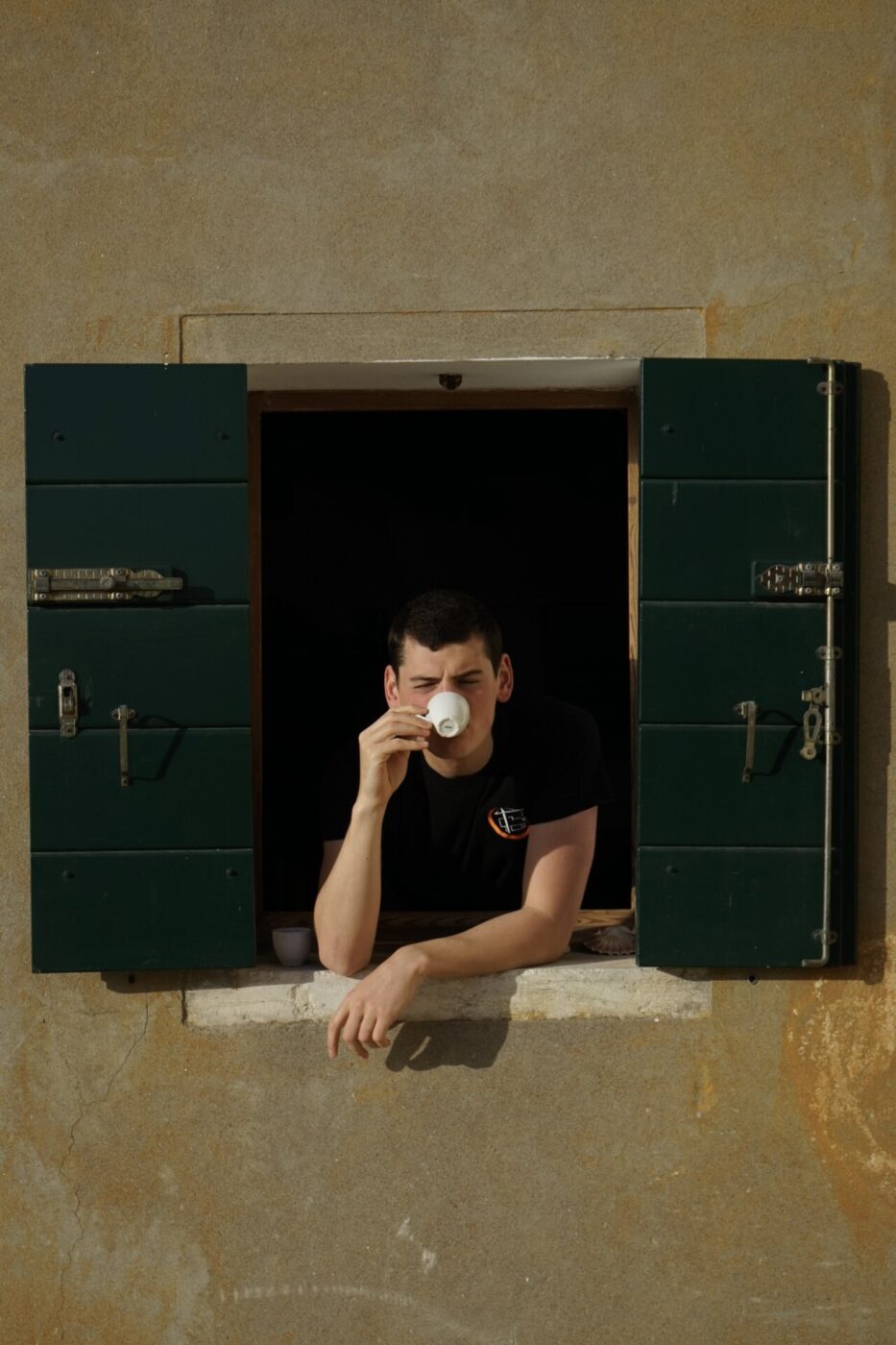a young man sipping an espresso between dark green shutters on Pellestrina