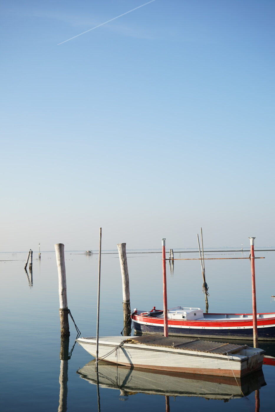 two small boats tied up between posts on perfectly still water in Pellestrina