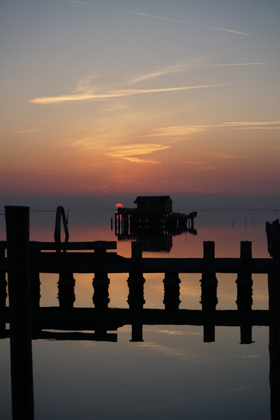 a hot pink moon and bright sunset over still, tranquil water with the reflection of raised houses and docks in Pellestrina