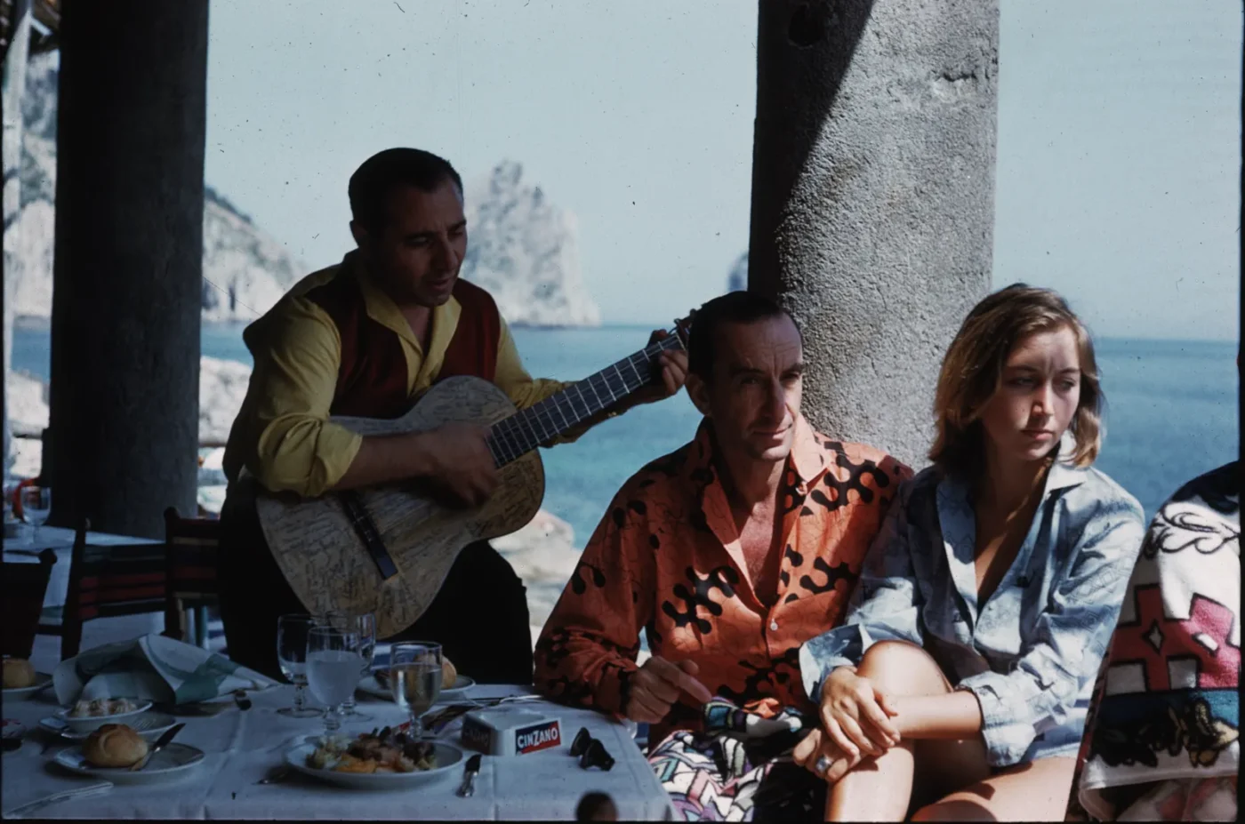 a photo of Capri in 1959 of three people sitting at a table on the water and one has a guitar