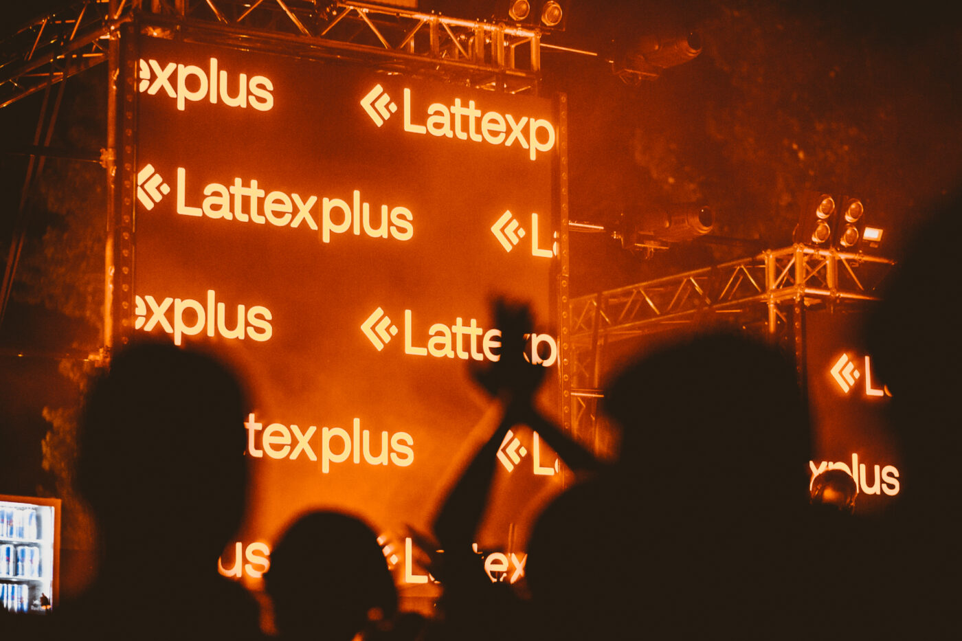 an outdoor event with a stage that says lattexplus in orange and has the dark silhouettes of people