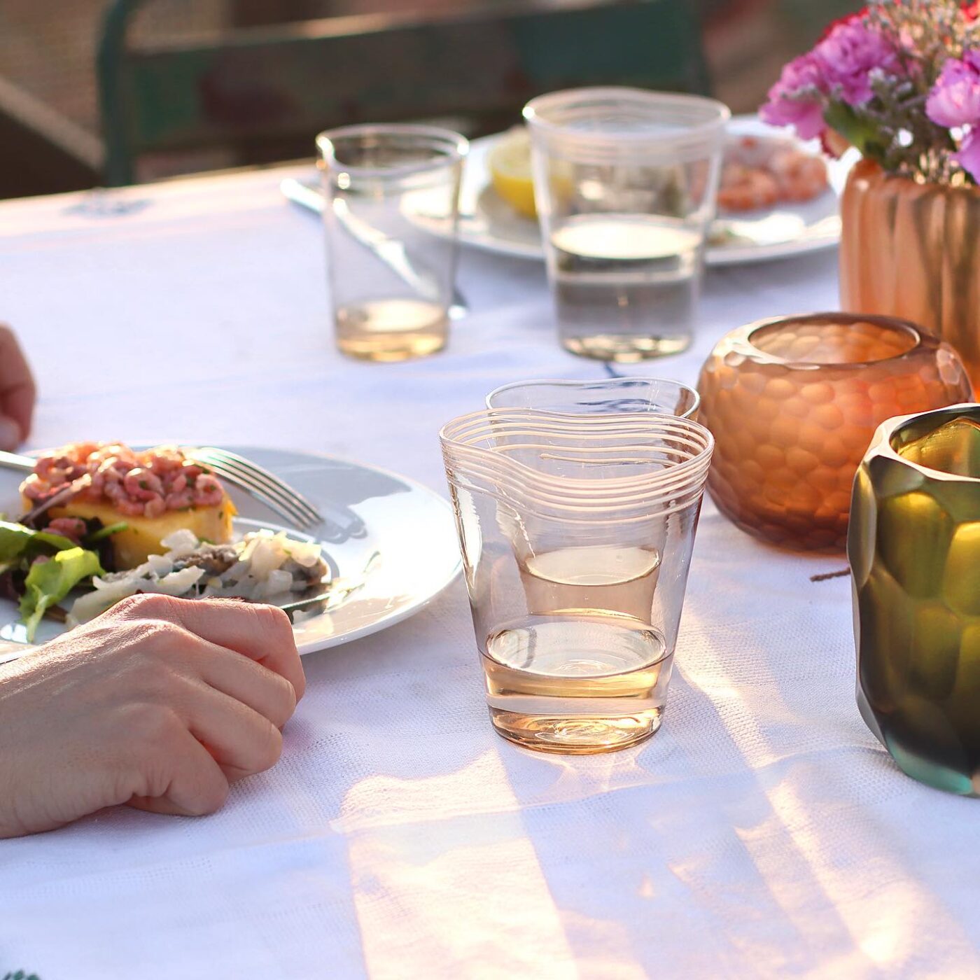 a photo of people sitting outside at a nice, set table with plates, flowers, and cups by Micheluzzi Glass