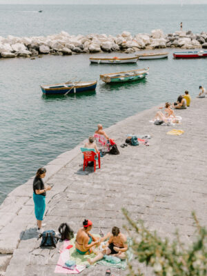the seafront in Naples with people laying on the concrete pathway and a few wooden boats anchored in the sea