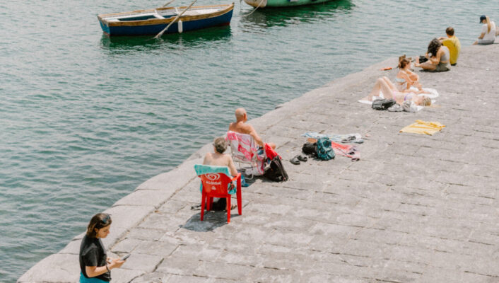 the seafront in Naples with people laying on the concrete pathway and a few wooden boats anchored in the sea