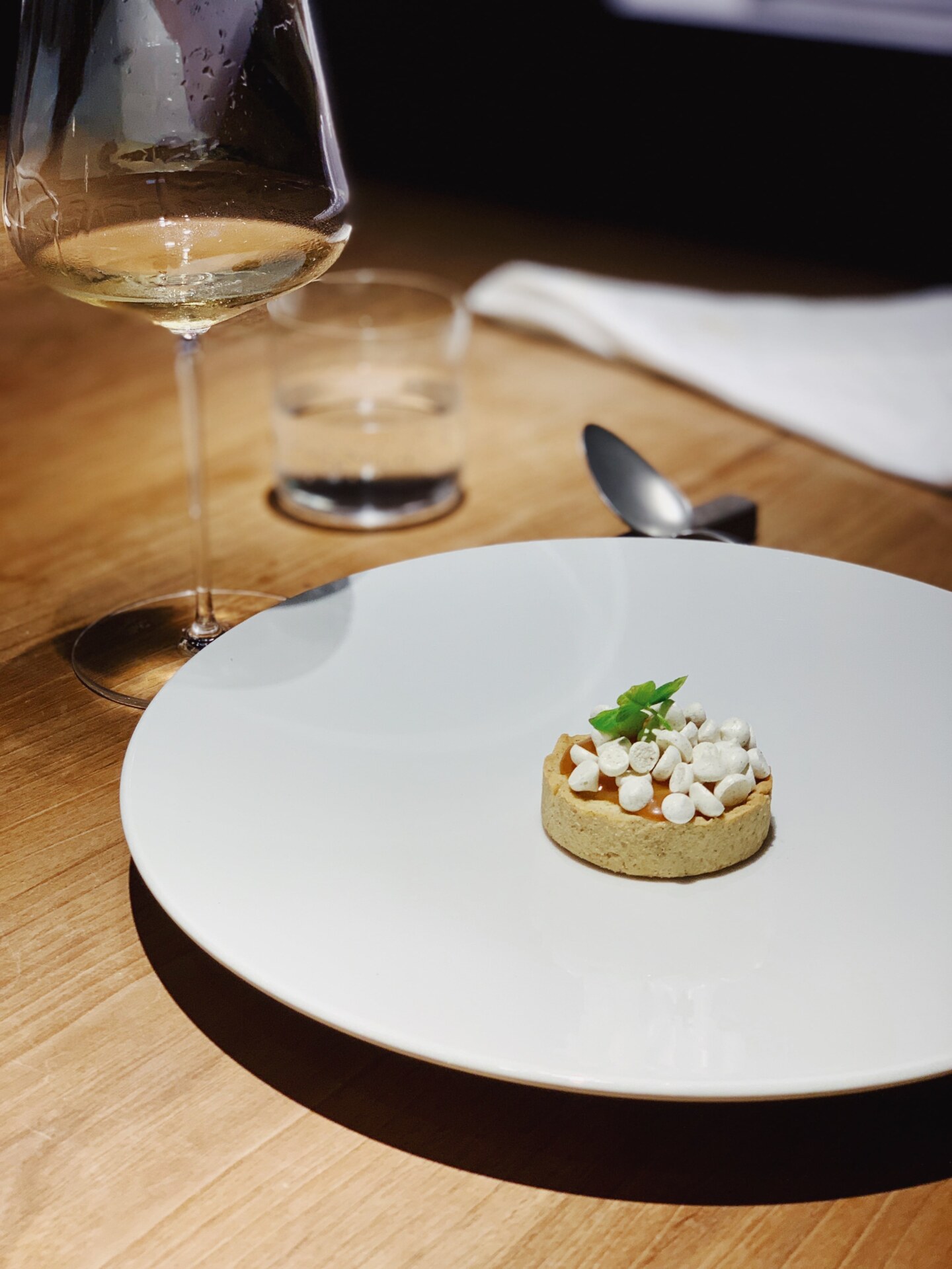 a tiny tart on a large white dish covered with small white chocolate chips next to a wine glass filled with white wine