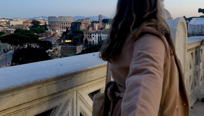 a woman shown from the back in a tan coat looking at the colosseum at dusk and holding the hand of someone behind her