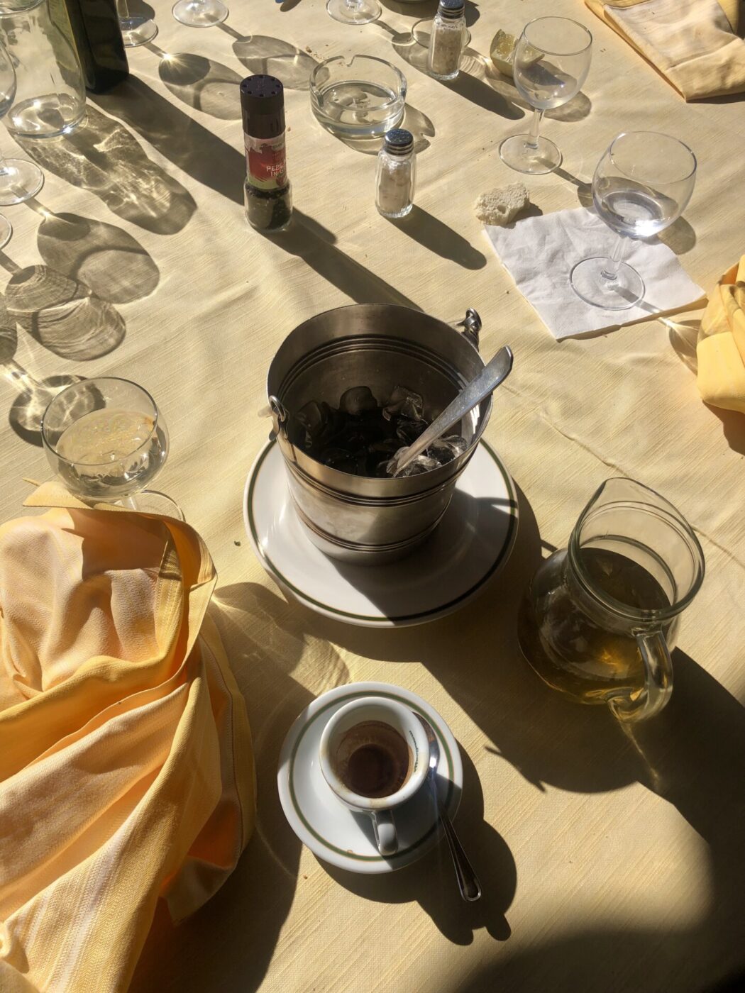 a table in the sun at a circolo with an ice bucket, pitcher of wine, espresso cup, and other empty cups and ash trays on a light yellow tablecloth